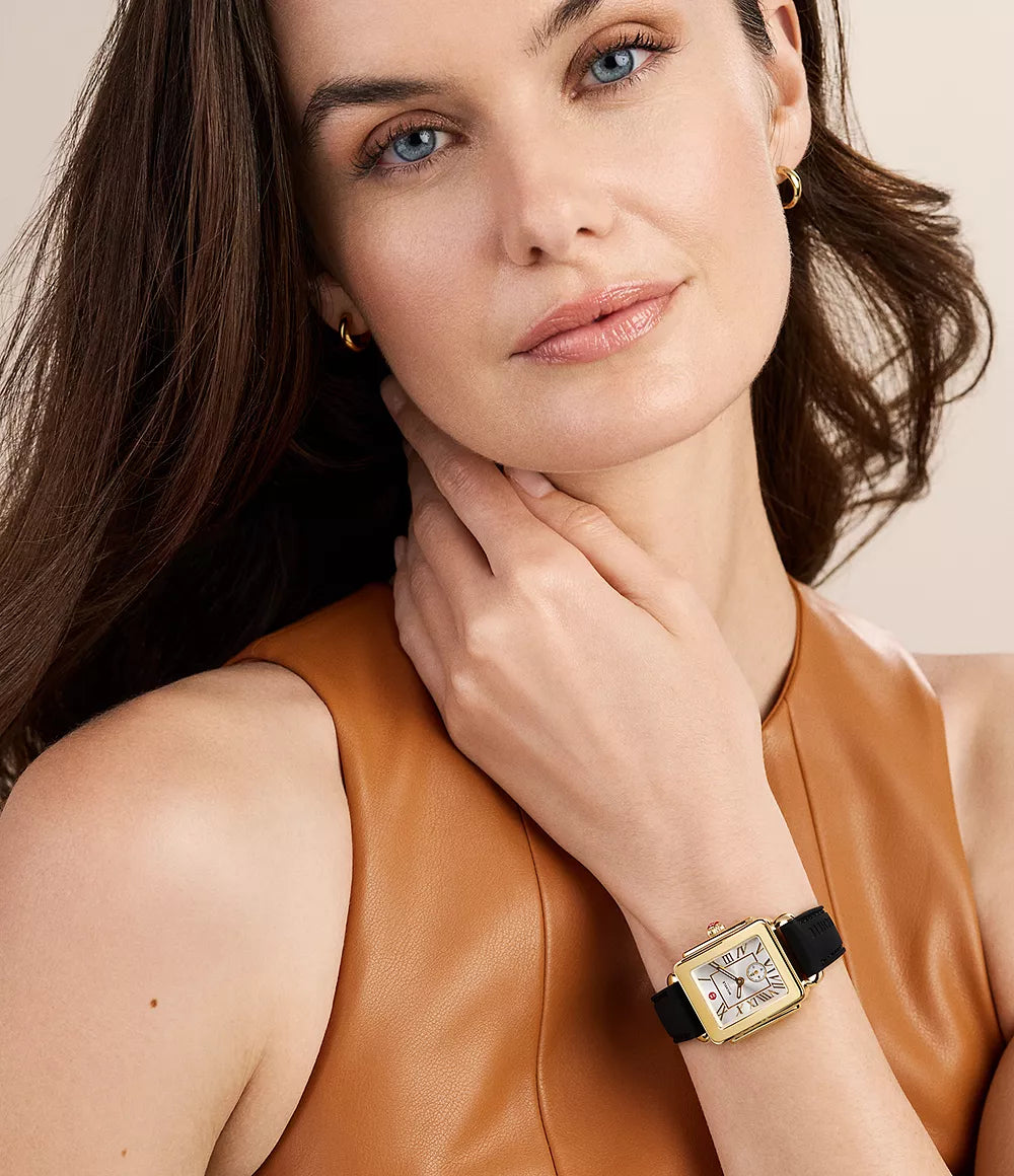 A woman wearing a gold-toned rectangular watch with a black strap and diamond-crystal face, paired with a tan leather top against a soft beige background.