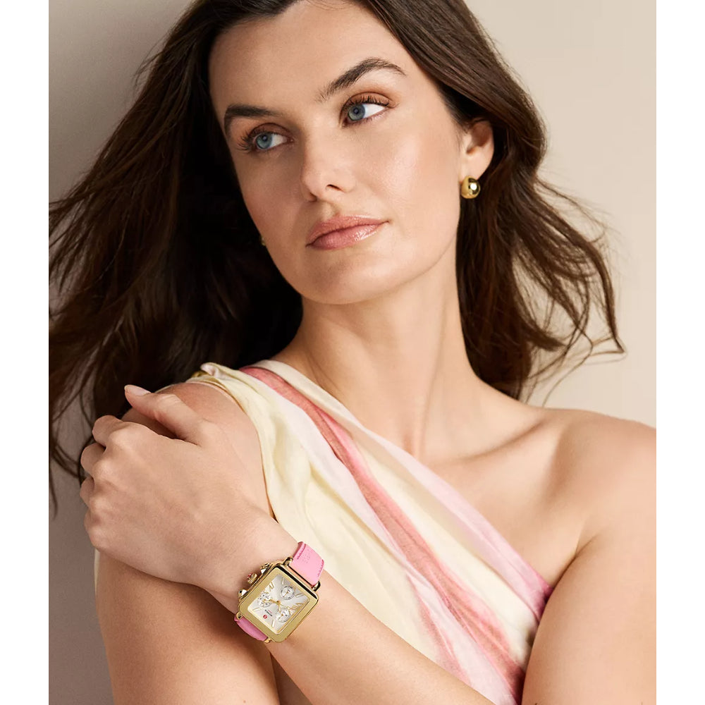 A woman with dark brown hair is shown in a close-up portrait wearing a gold-toned rectangular watch with a light pink strap. She is wearing a cream and pink draped top, and a small gold earring is visible.