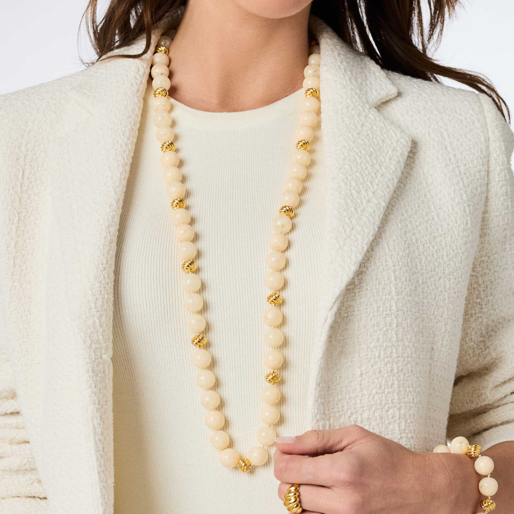 Person wearing a long beige beaded necklace with gold accents, paired with a white outfit and beige jacket.