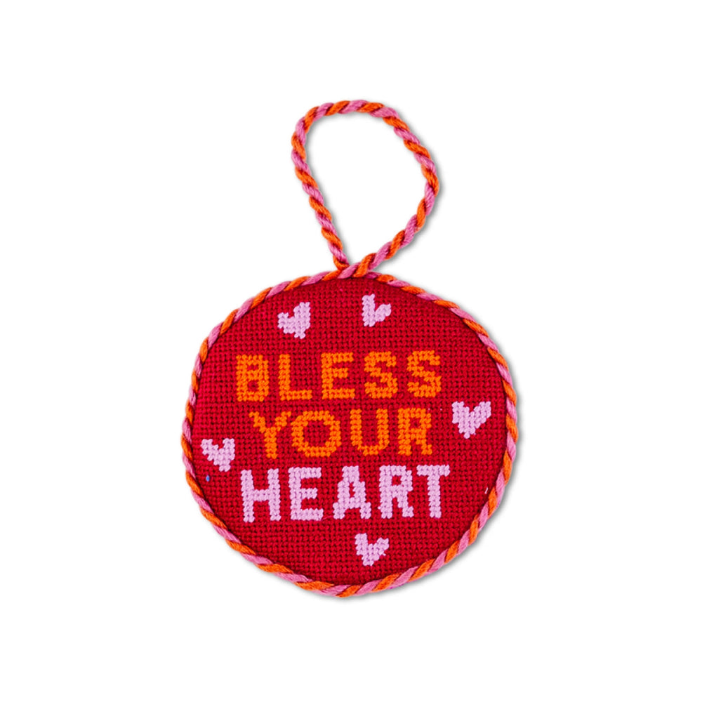 A round red needlepoint ornament with a hanging loop, featuring the words 'BLESS YOUR HEART' in orange, pink, and white lettering, surrounded by small pink hearts, on a white background.