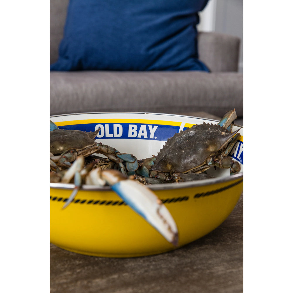 A side photo of a 4 qt yellow,white & blue bowl with an Old Bay seasoning label on a white background with live blue crabs in it on a wooden table