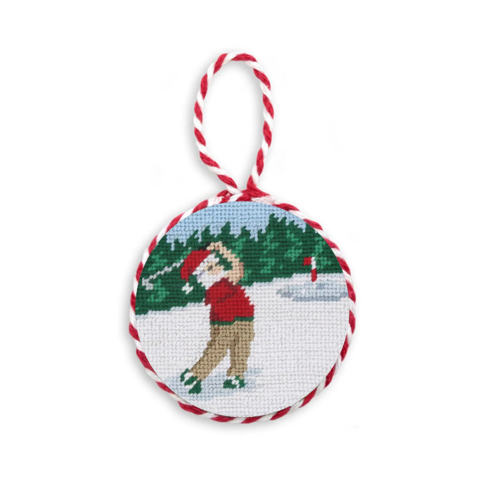A circular counted cross-stitch ornament featuring Santa Claus swinging a gold club on a snowy background with green trees, hanging from a red and white twisted cord loop.