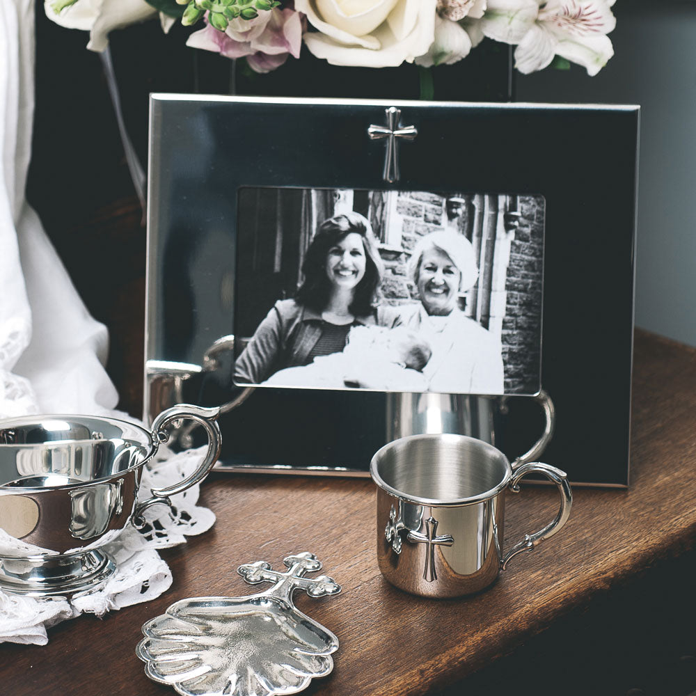 Horizontal silver photo frame with a cross on the top on a table with a silver cup with a cross and other silver cups and items with a bouquet of pink flowers.