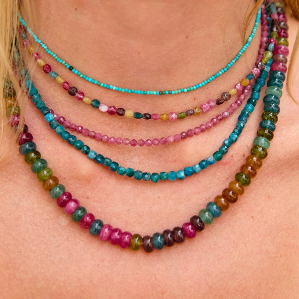 A close-up of a person wearing five layered beaded necklaces in various vibrant gemstone colors including turquoise, pink, and the last one is multicolor