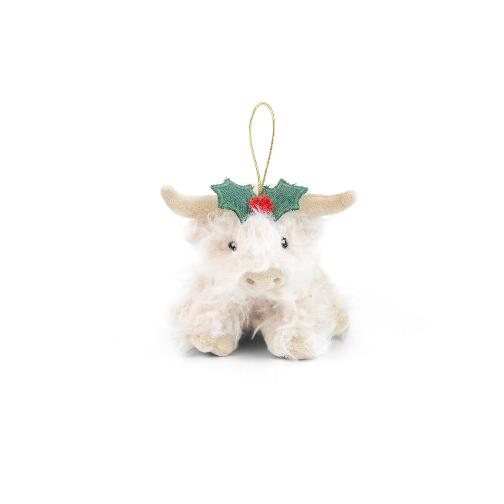 A small plush Highland cow Christmas ornament with cream-colored wool, tan horns, a green felt holly headpiece with a red berry accent, and a gold hanging loop, shown on a plain white background.