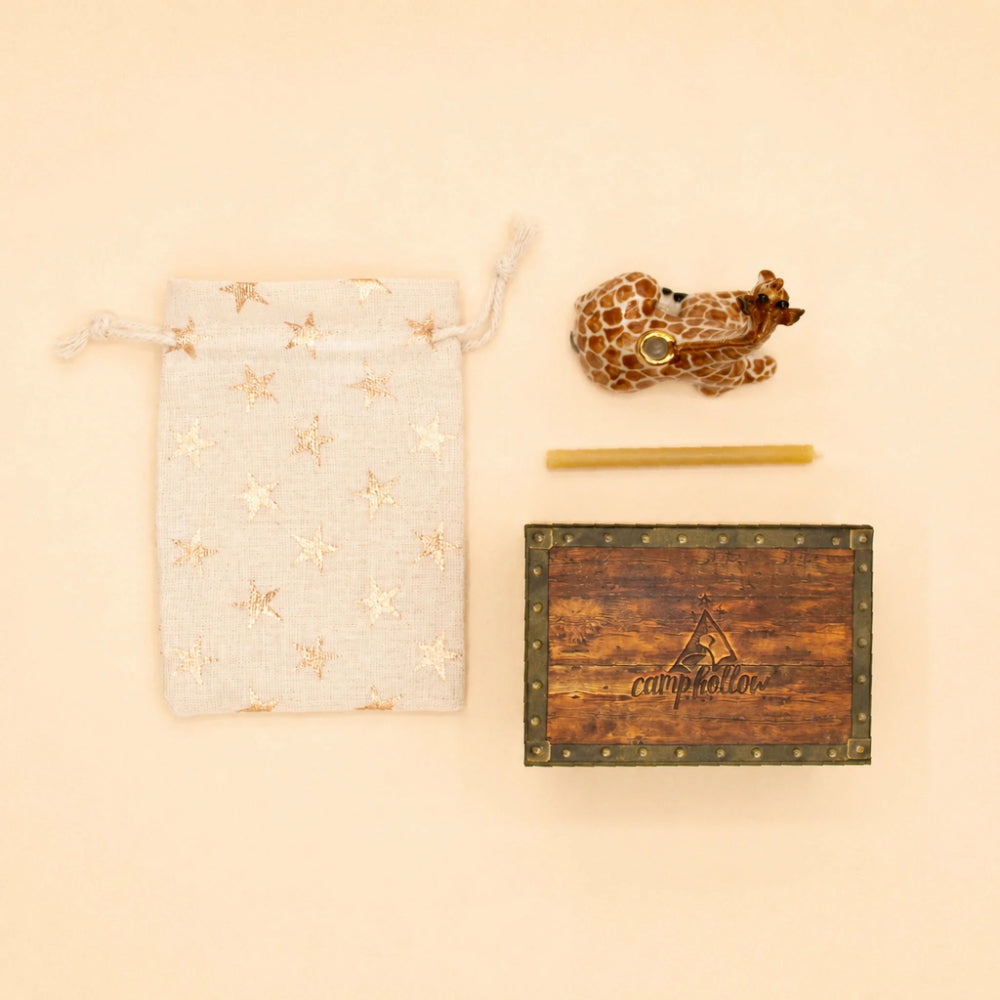 A giraffe-themed gift set on a warm beige background, featuring a cream drawstring pouch with gold star patterns, a small carved giraffe figurine, a yellow candle, and a rustic wooden box engraved with 'campmation'.