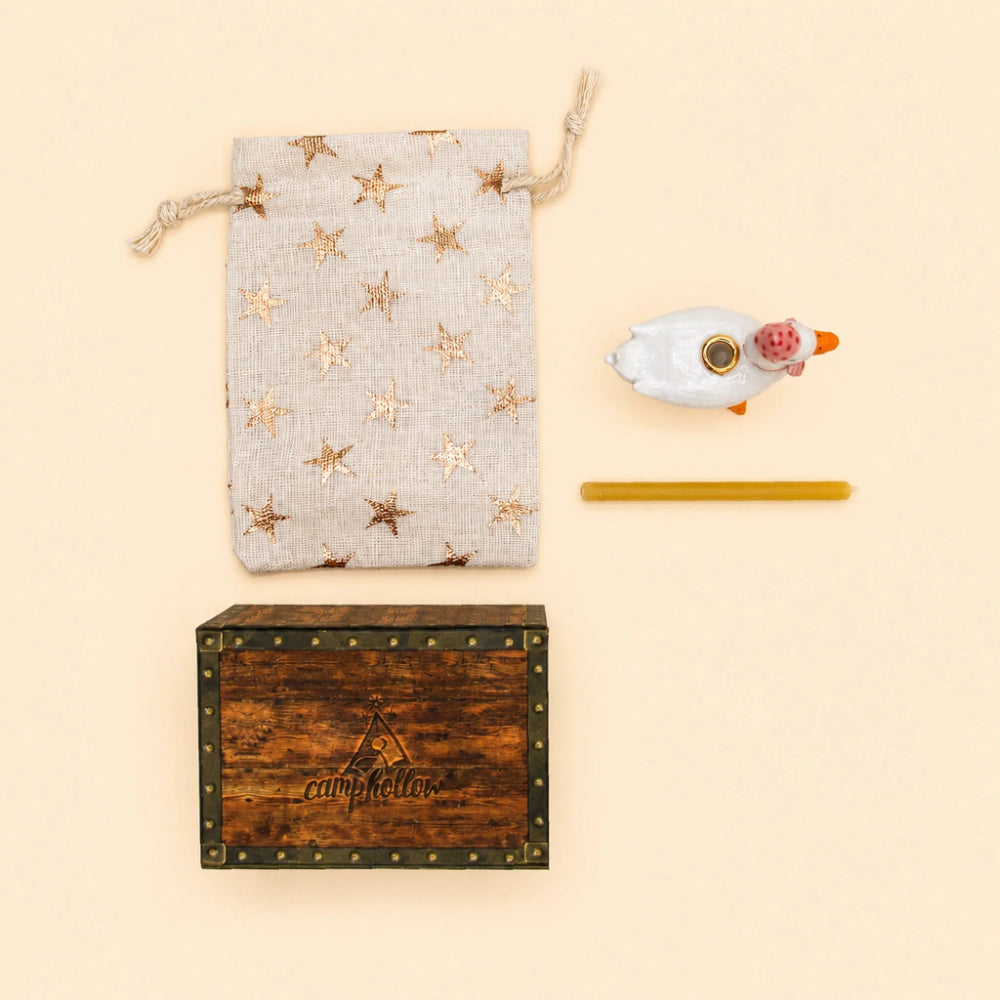A curated gift set on a warm cream background, featuring a beige linen drawstring pouch with metallic gold star patterns, a rustic dark brown wooden box with brass corner studs and 'campkeller' branding, a small white duck figurine with a pink beak, and a yellow glass straw.