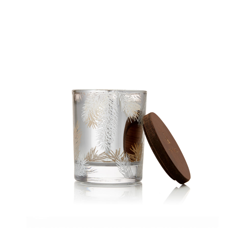A clear glass candle jar with etched silver pine branches, shown next to its dark brown wooden lid on a white background.