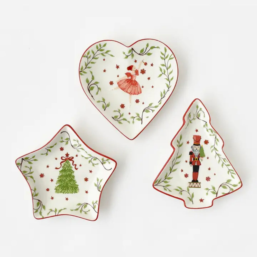 Three ceramic Christmas-themed dishware pieces on a white background: a heart-shaped dish with a red dress illustration, a star-shaped dish with a green Christmas tree, and a tree-shaped dish with a nutcracker.