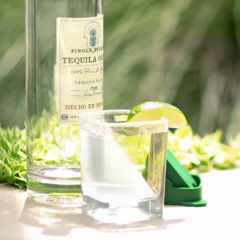 Clear glass tumbler with green tequila triangle lid with a tequila bottle against a blurred outdoor setting.