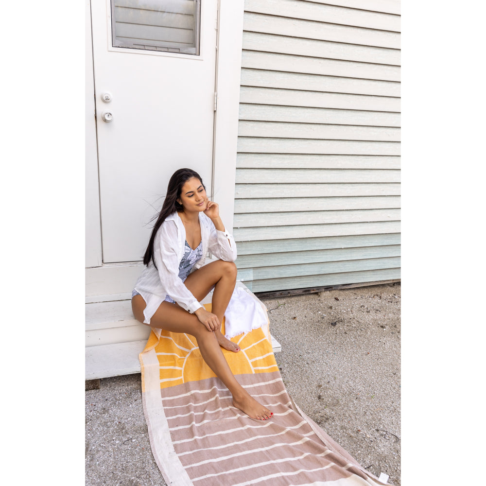 Woman sitting on a beach towel that has a sun and wave design in yellow and brown by a white building at the beach.