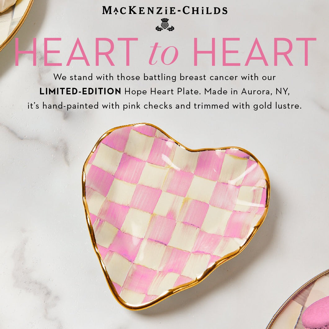 A heart-shaped MacKenzie-Childs plate with pink checks and gold lustre trim on the edge, with the brand's tagline 'We stand with those battling breast cancer'.