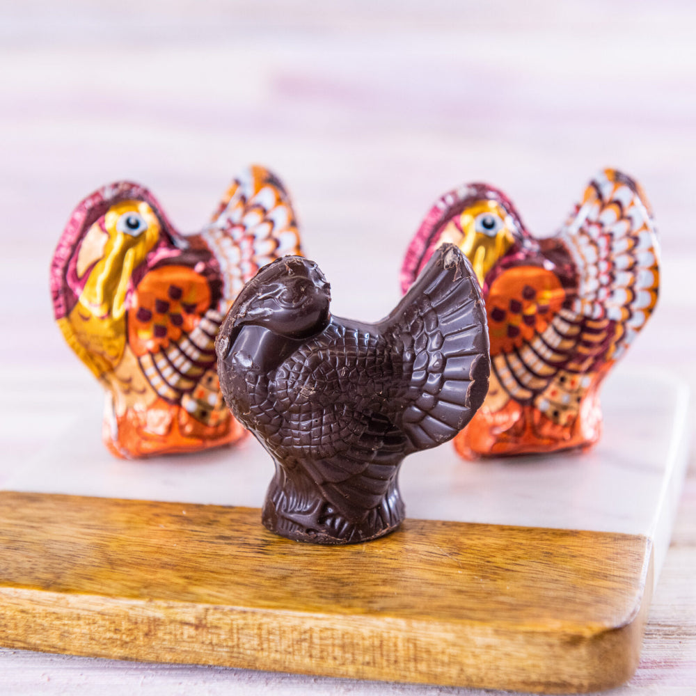 A dark brown chocolate turkey figurine in the foreground, with two colorful foil-tinted turkey chocolates with packaging behind it on a light surface and a wooden board.