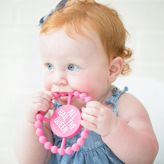 Baby holding a pink teething ring with text, wearing a blue dress against a white background