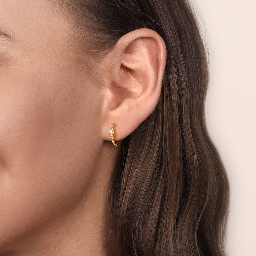 Close-up of an ear wearing a gold hoop earring with a small gemstone on a neutral background