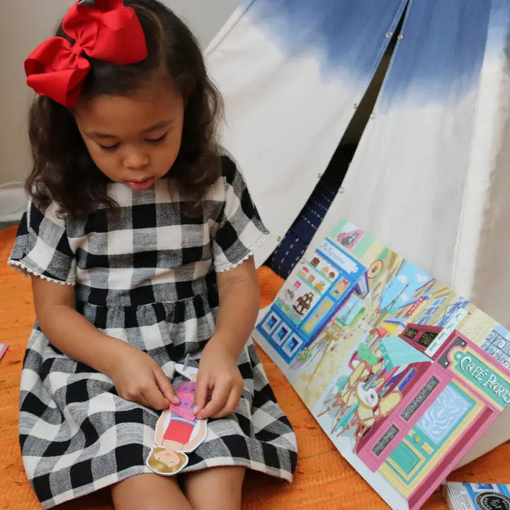 Child in a checkered dress with a red bow, sitting on the floor with colorful paper doll set.