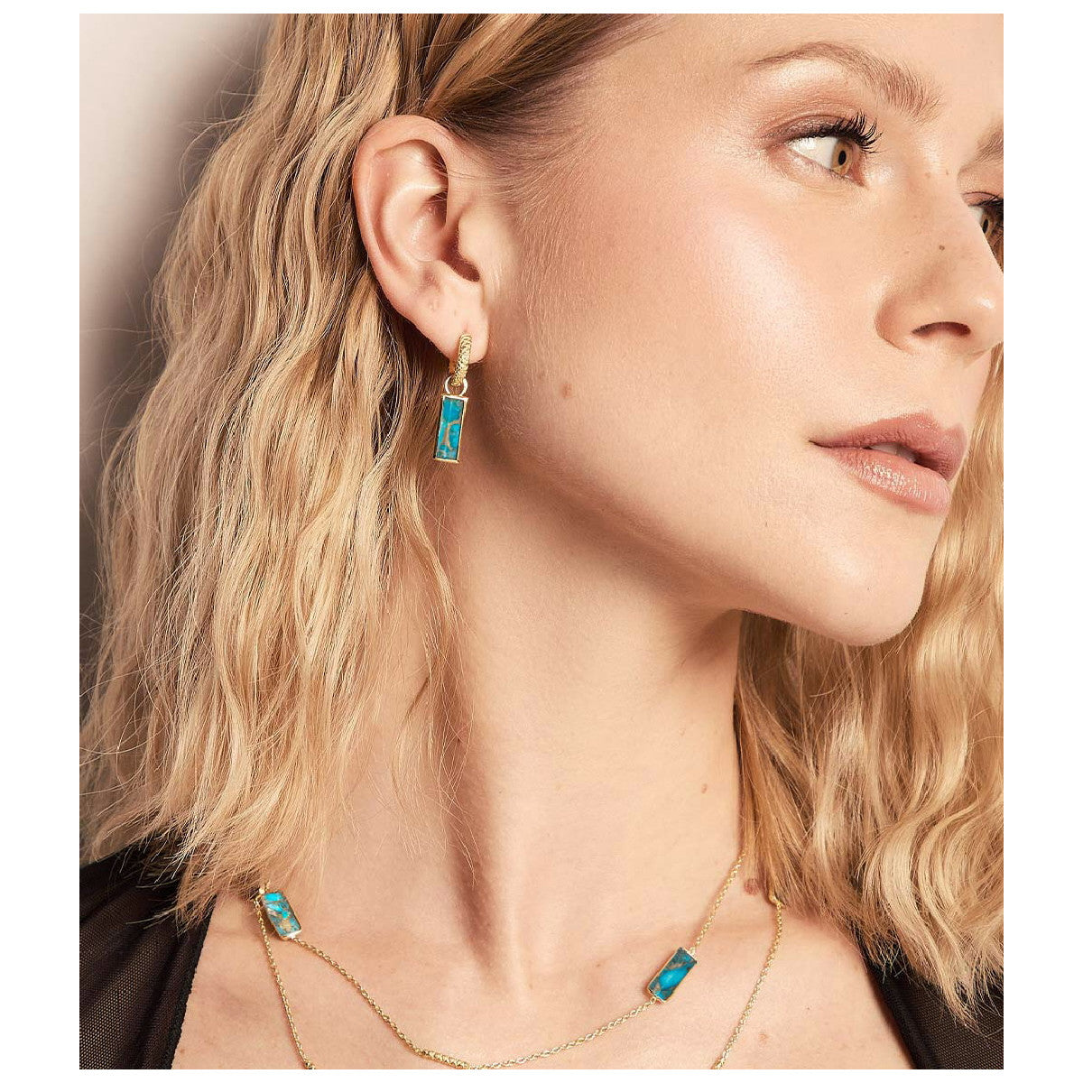 A close-up of a woman wearing a gold necklace and matching drop earrings with rectangular turquoise-blue stones, shown against a soft beige background.