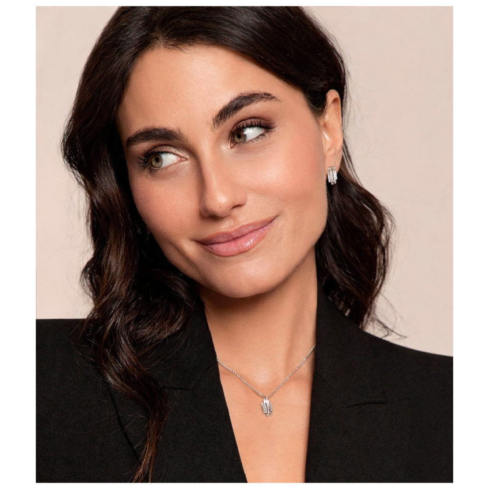 A woman with dark wavy hair wearing a silver pendant necklace and matching stud earrings, dressed in a black blazer against a soft beige background.