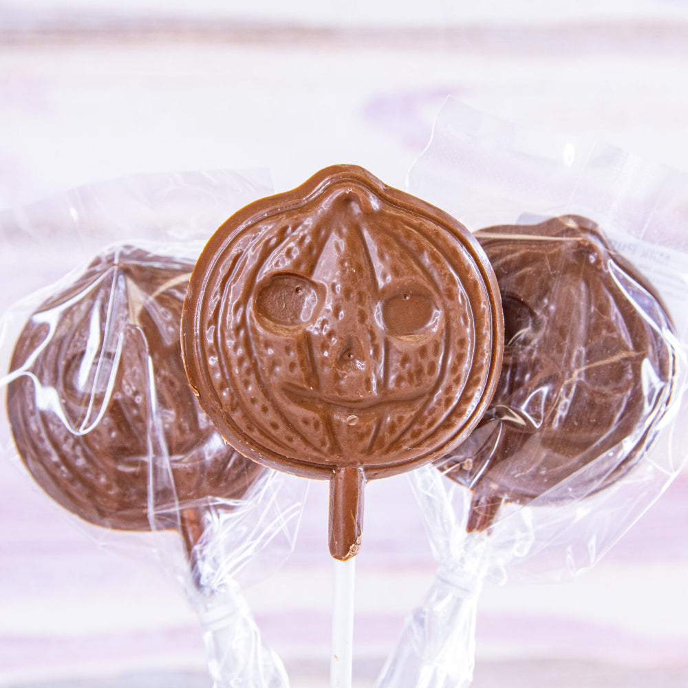 Three pumpkin-shaped milk chocolate lollipops on sticks, wrapped in clear cellophane packaging against a soft, light pink and white background.