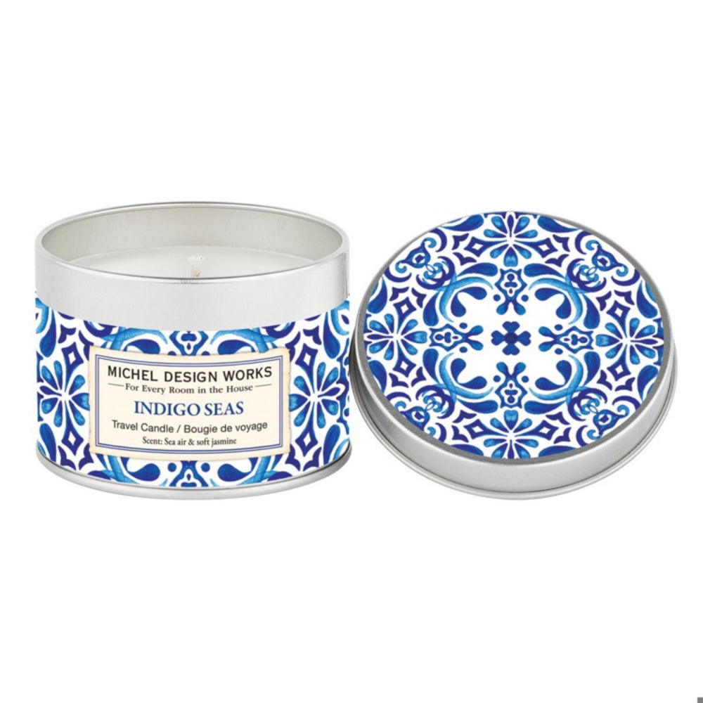A travel candle from Michel Design Works, with a blue and white decorative lid and a label that reads 'Indigo Seas'. The candle is shown with its lid open, revealing the ocean-themed design.