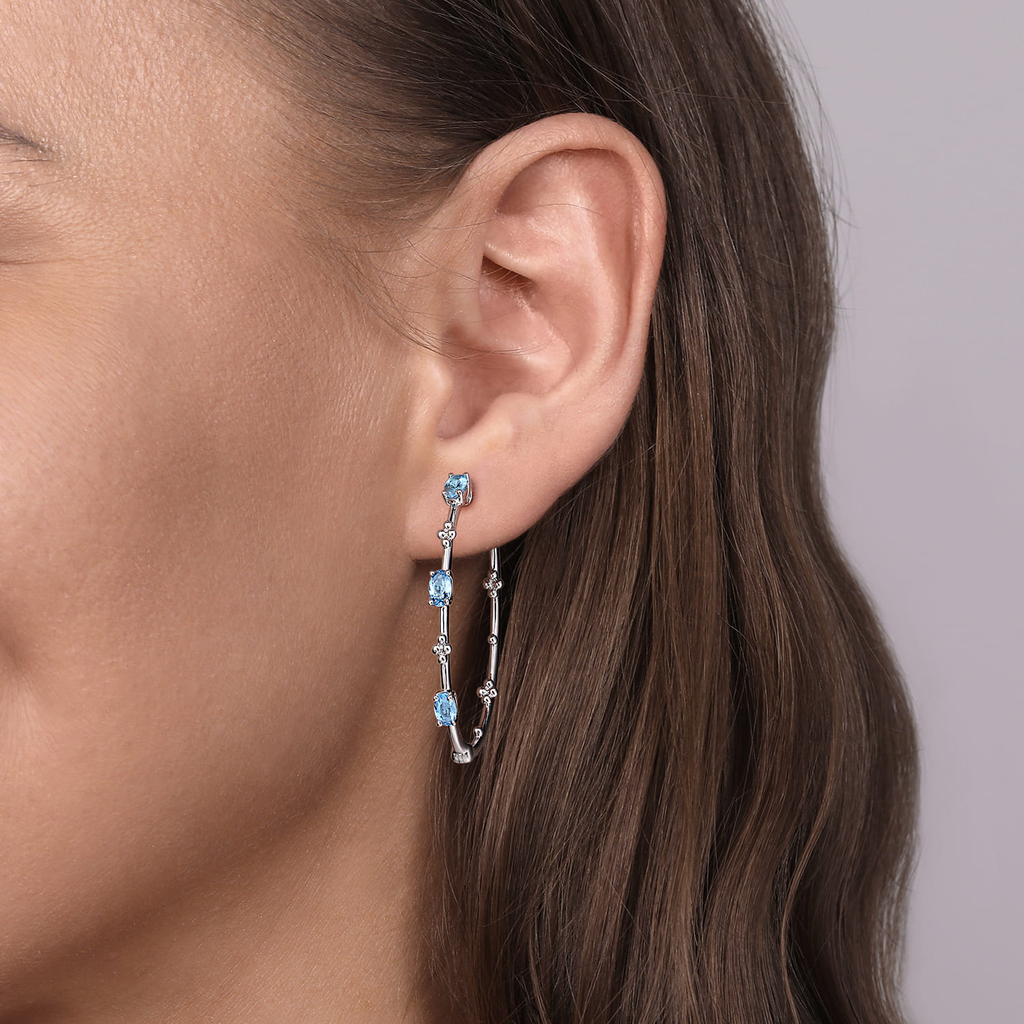 Close-up of a woman's ear wearing a silver earring with blue gemstones against a neutral background