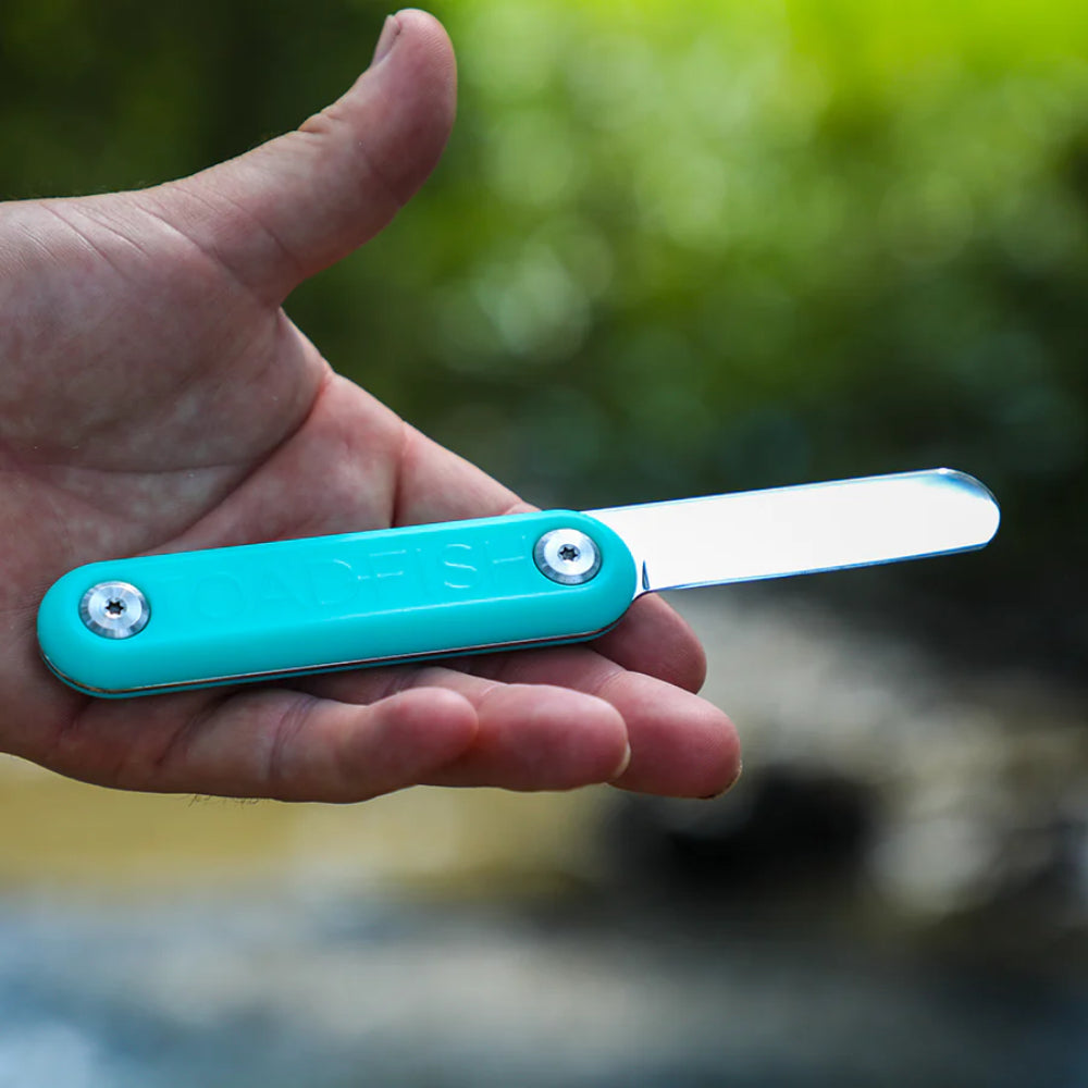 A hand holding a turquoise clam knife with a black metallic blade on a blurred outdoor backround.