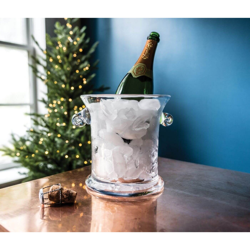 Clear glass ice bucket with glass handles with a wine bottle with ice in it, on a brown wood table in a blue painted room with a christmas tree with white lights on it next to a window