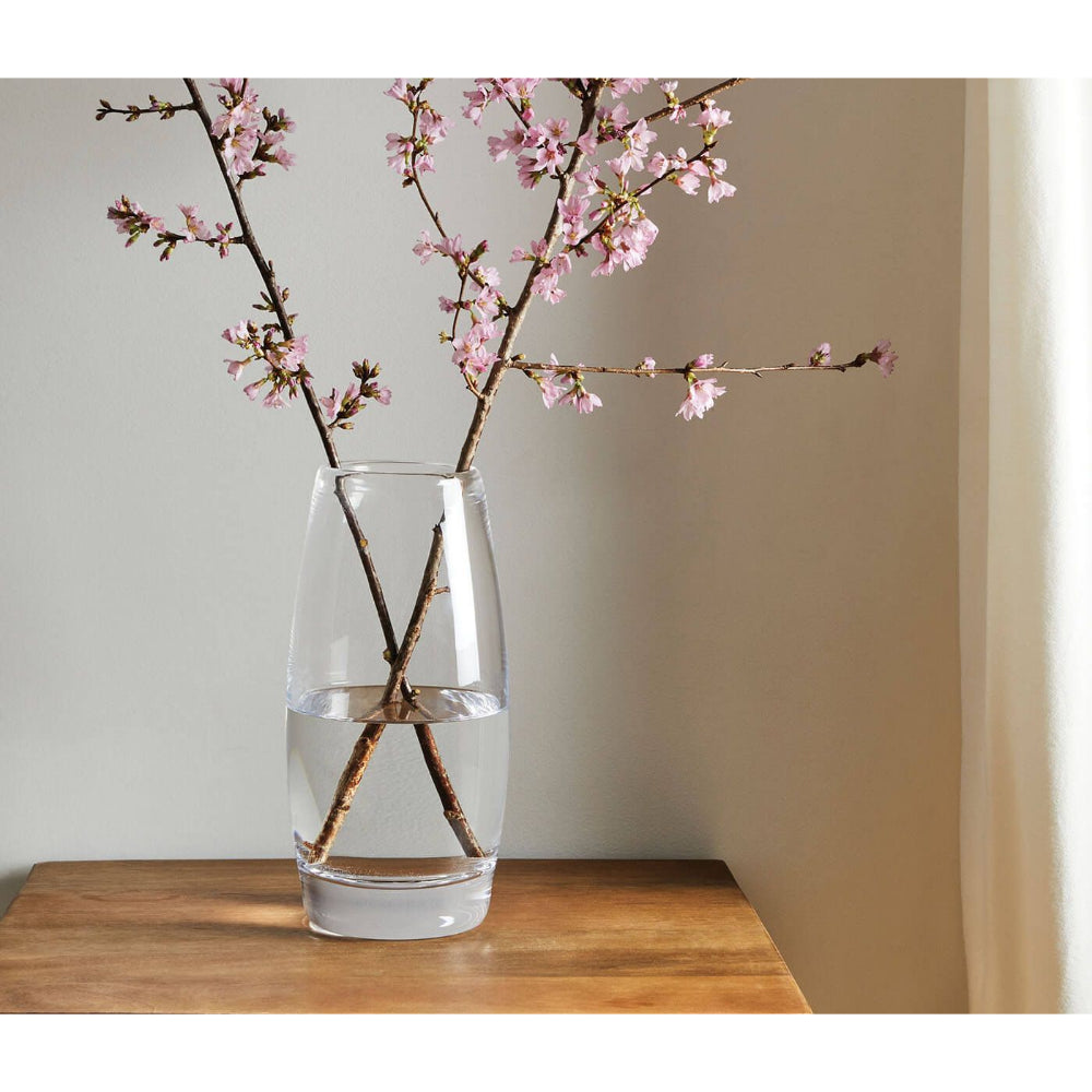 A clear glass vase with pink flowers in it on a wood table with a white painted wall next to a window with white blinds