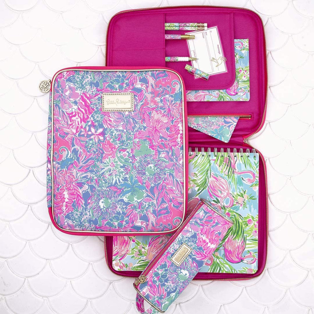 A anged side photo of a Lilly Pulitzer pencil pouch with a multicolored floral pattern and gold pu accents, featuring a zipper closure and a gold Lilly logo charm zipper pull with a note book and profolio in the same floral pattern.