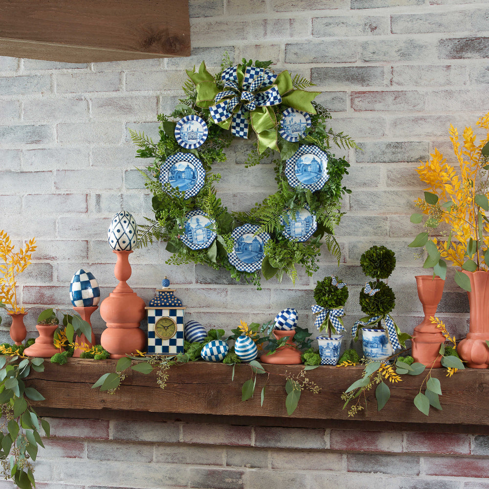 An life style photo of a decorative clock with royal blue and white checkered design on a mental with a holiday weath with other holiday decor in royal blue and white checks.