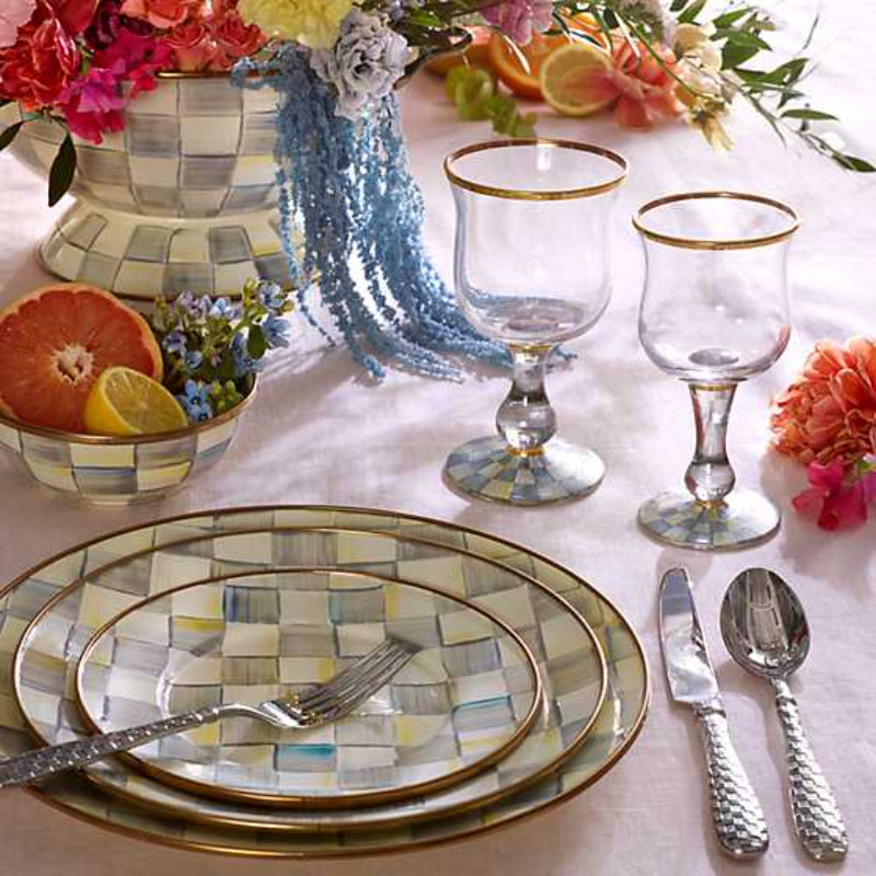 Clear glass with gold rim and decorative base royal blue checkard base on a table with a white cover with grey checkard plates and other tableware and grey checkard vase with flowers and fruit