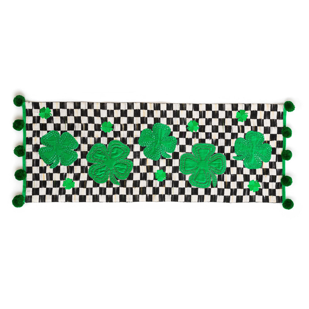 Black checkered table runner with green shamrock designs and pom-pom edges on a white background
