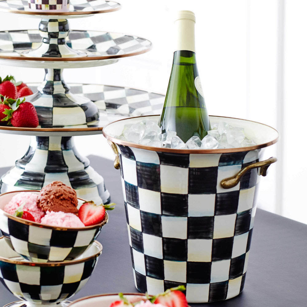A hand-painted, glossy, black and white checkered enamel wine cooler with bronzed stainless steel rim and handles on a table with wine and ice in it with other simular items.