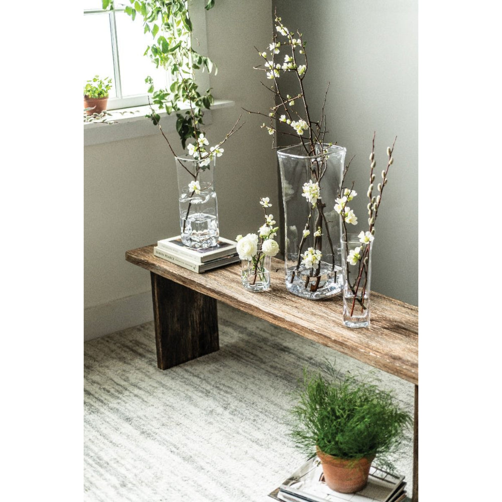 Clear glass vases on a bench with plants with white flowers on it in a white colored room with wood floor