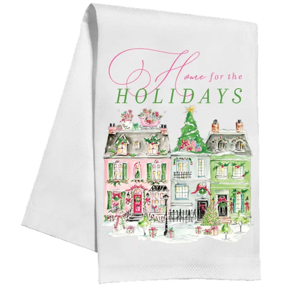 A white kitchen tea towel featuring a festive holiday illustration of three colorful houses, a Christmas tree, and the text 'Home for the HOLIDAYS' in pink and green script.