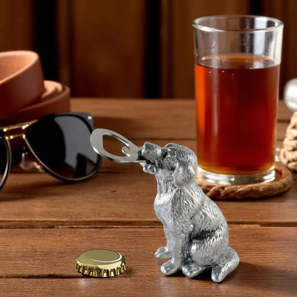 A silver dog-shaped bottle opener resting on a wooden table next to a gold bottle cap, with a glass of amber liquid, sunglasses, and a brown belt in the background.