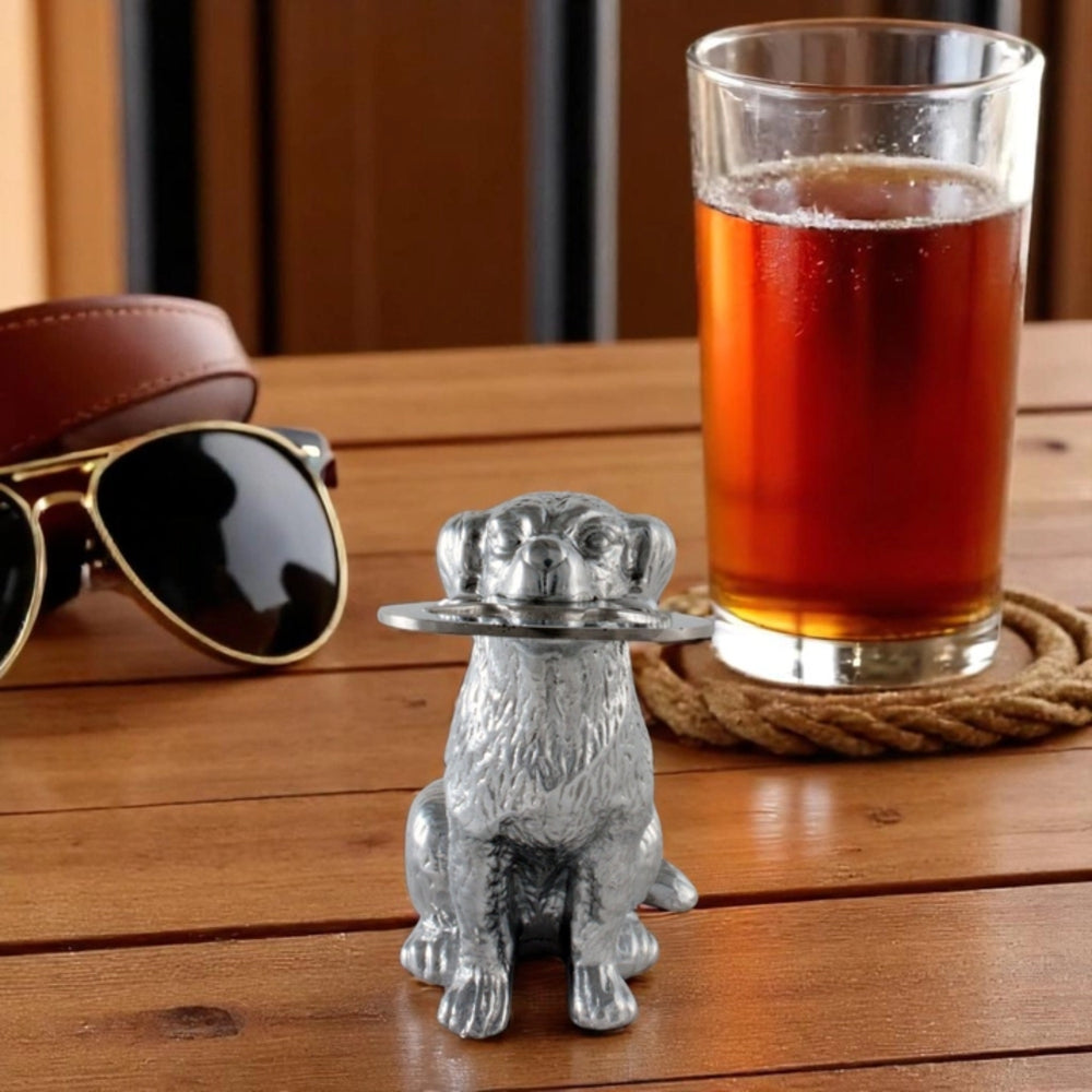 A small metallic silver dog figurine holding a glass in its mouth, placed on a wooden tabletop with sunglasses, a brown leather strap, and a glass of amber liquid on a rope coaster in the background.