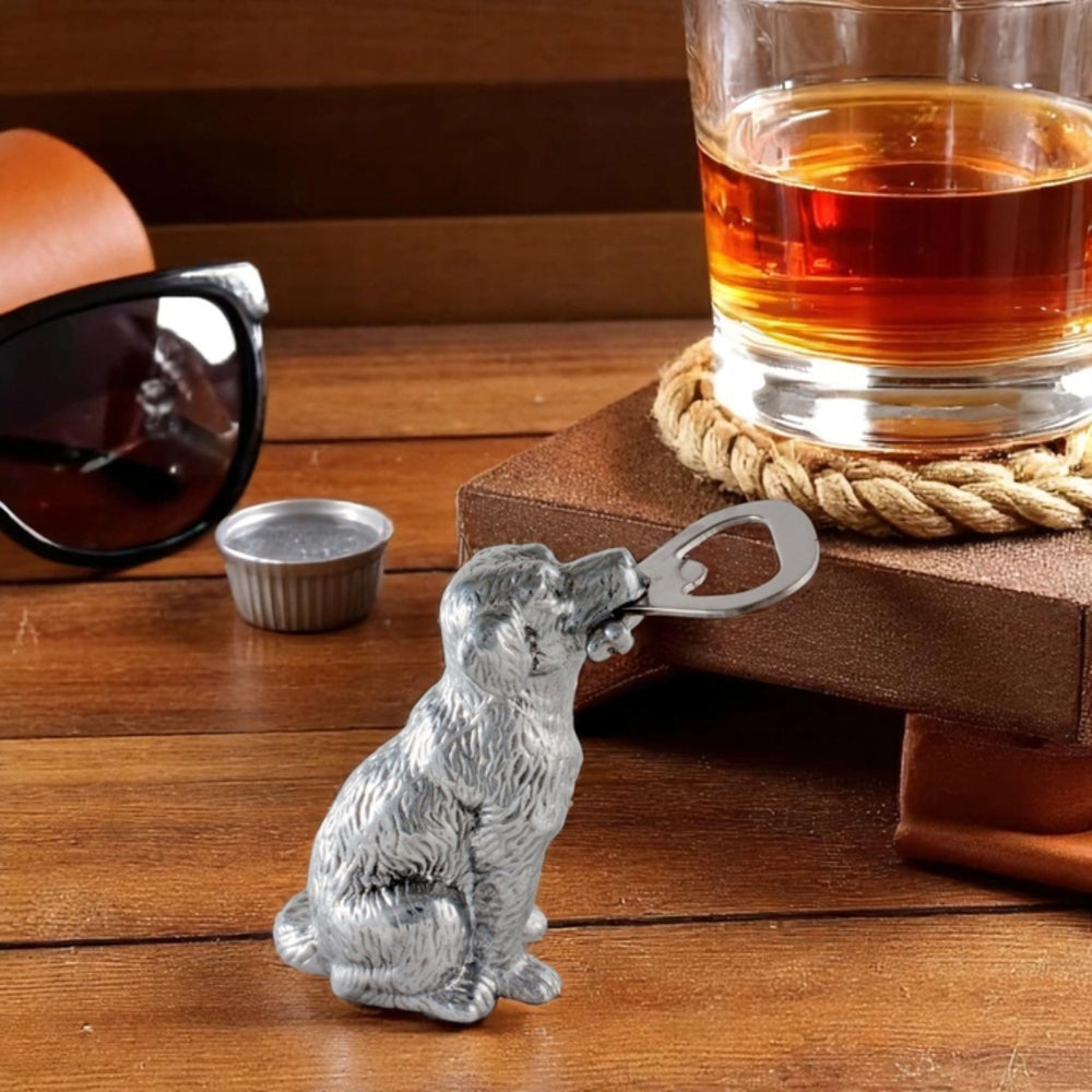 A silver metal dog-shaped bottle opener resting on a wooden table, styled with sunglasses, a small metal cup, a glass of amber liquid on a wooden block, and a rope coaster.
