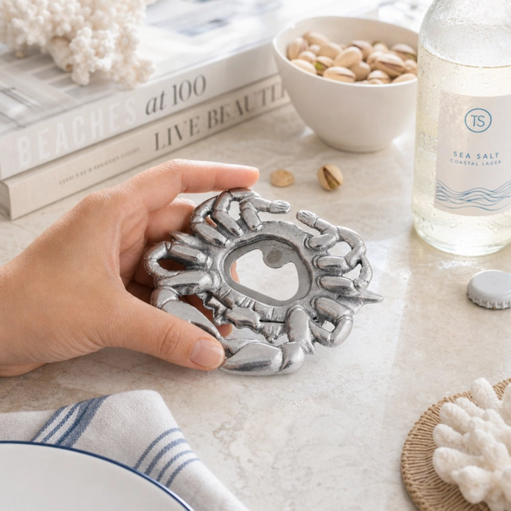 A hand holds a silver lobster-shaped bottle opener on a light marble tabletop, surrounded by nautical-themed props including a bottle of sea salt, books, and a bowl of pistachios.