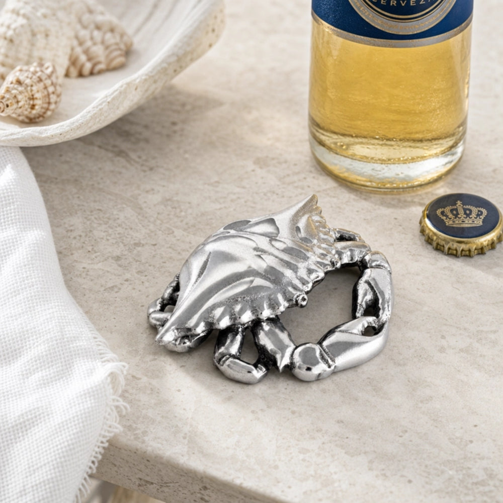 A silver crab-shaped bottle opener resting on a light beige stone surface, styled with a bottle cap, a decorative dish, and a glass of beer nearby.