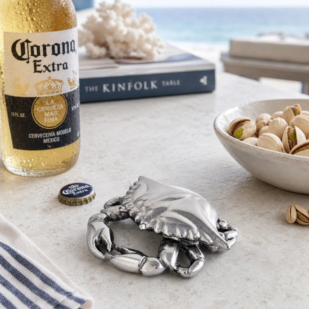 A silver-toned metal crab-shaped bottle opener resting on a light stone surface, styled with a Corona Extra beer bottle, a cap, a bowl of pistachios, and a book in a beach-side setting.