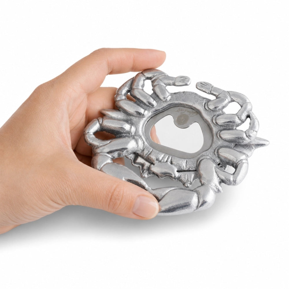 A hand holding a silver metal lobster claw bottle opener against a plain white background.