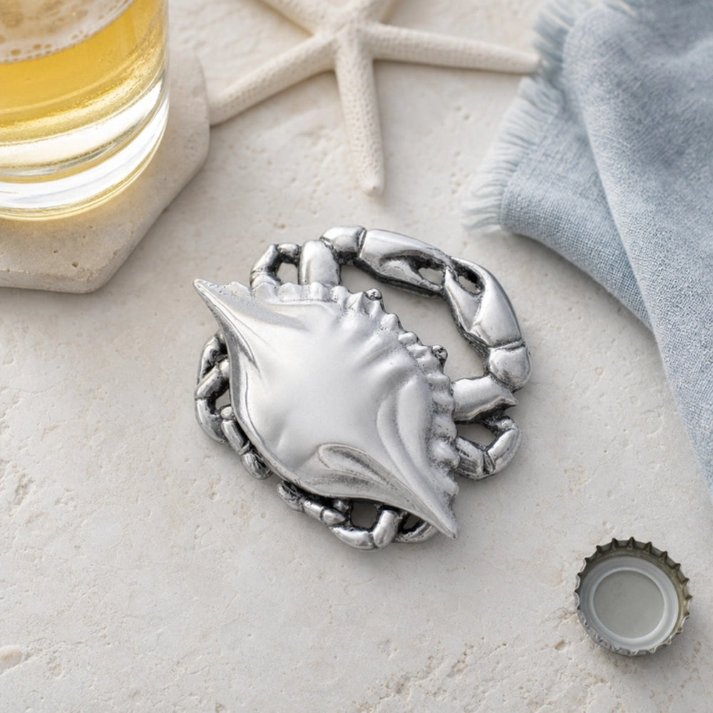 A silver metal lobster-shaped bottle opener styled on a light beige textured surface with beach-themed props including a starfish, a glass of beer, a blue towel, and a bottle cap.