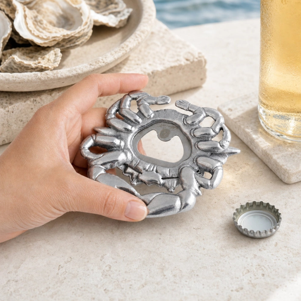 A hand holds a silver metal crab-shaped bottle opener on a light stone surface, with a bottle cap nearby and oyster shells in the background.