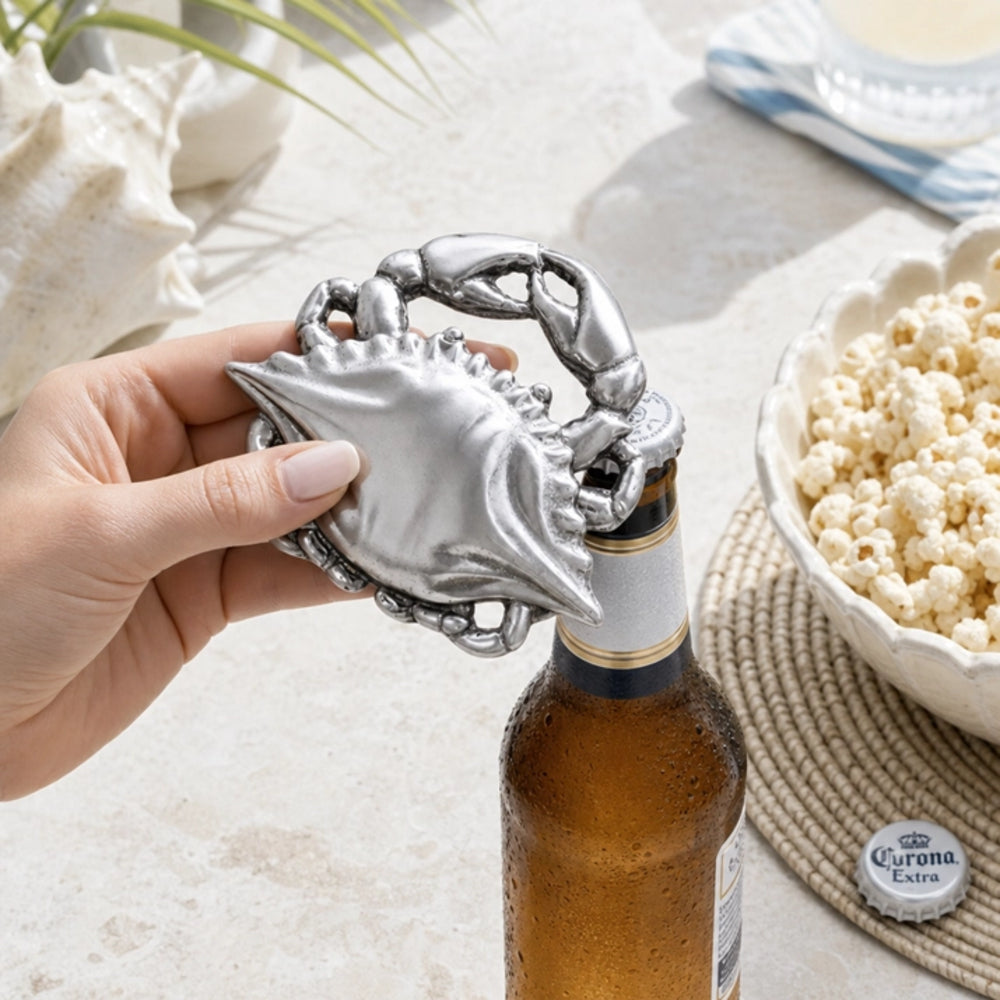 A hand holds a silver lobster-shaped bottle opener attached to a brown beer bottle, with a bowl of popcorn and a Corona Extra cap nearby on a light stone surface.