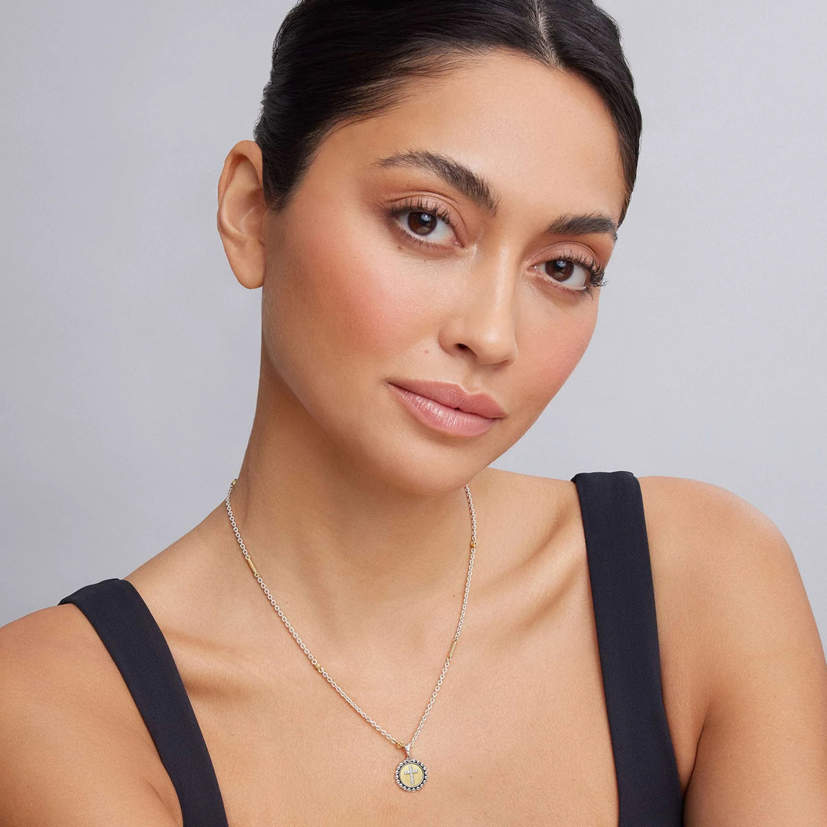 A close-up of a woman wearing a delicate gold chain necklace with a circular pendant featuring a light blue and white pattern, paired with a black top against a neutral gray background.