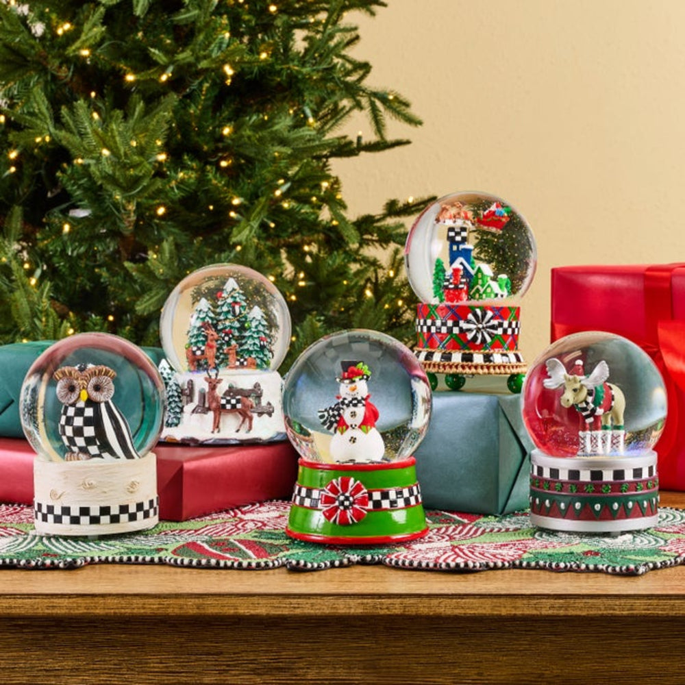 Five decorative snow globes with holiday-themed designs are arranged on a table runner in front of a Christmas tree and wrapped gift boxes. The globes feature winter scenes and characters, with colorful bases in red, green, black, and white.