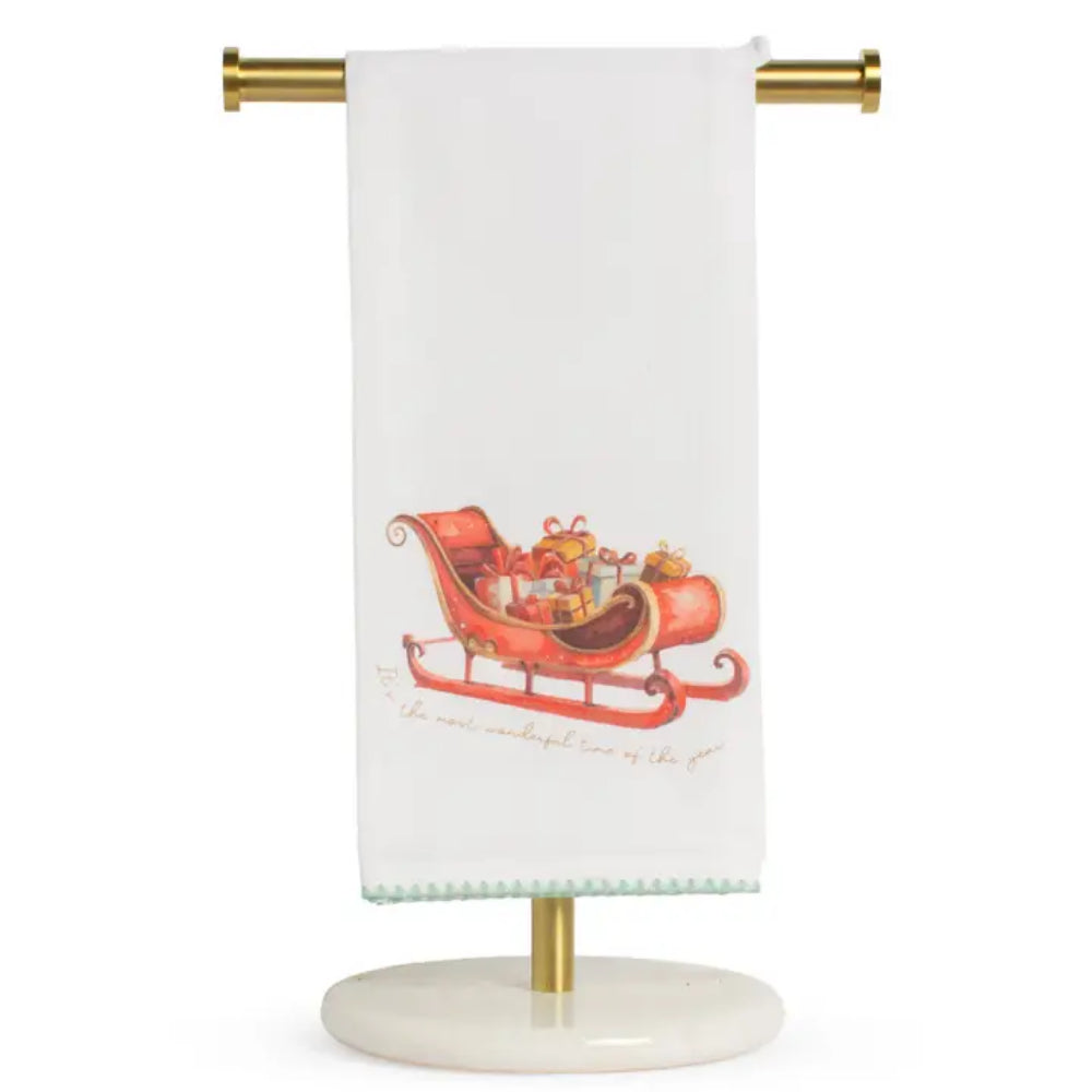 A white kitchen towel hanging from a gold-colored stand, featuring an illustration of Santa's sleigh filled with presents and the phrase 'The most wonderful time of the year' in cursive.
