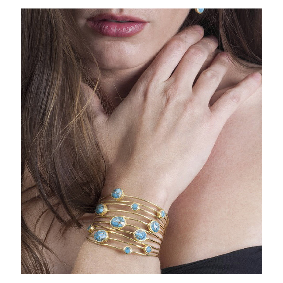 A close-up of a woman wearing a stack of gold cuff bracelets, each adorned with multiple round light blue gemstones, resting her hand near her neck.