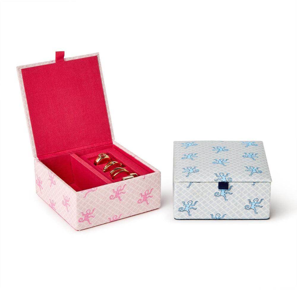 Two decorative jewelry boxes on a white background: one open pink box with a red interior holding gold rings, and one closed white box with blue patterns and a small blue clasp.